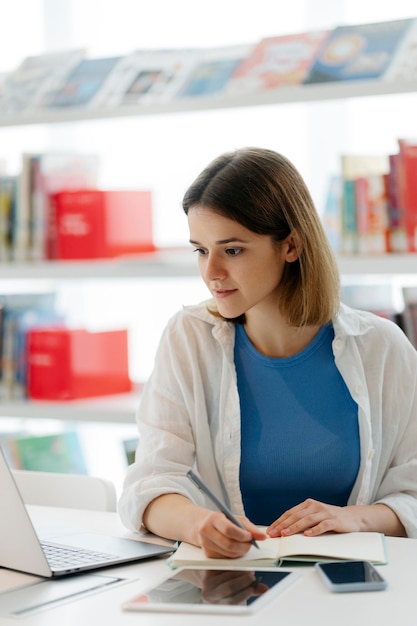 Estudiante que estudia usando una computadora portátil tomando notas preparación para el examen en la biblioteca Concepto de educación en línea