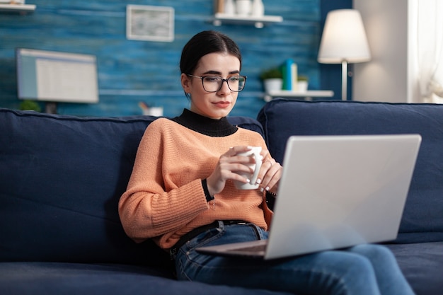 Foto estudiante que mira la computadora portátil durante los cursos de administración en línea sentado cómodo en el sofá en la sala de estar. mujer leyendo la lección de marketing en la plataforma universitaria, adolescente estudiando a distancia