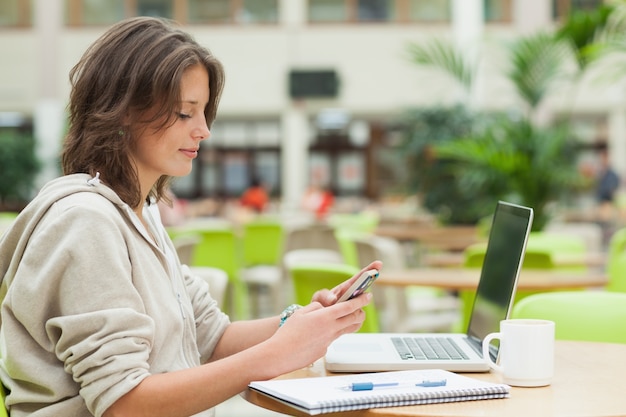 Estudiante que usa el teléfono celular y la computadora portátil en la tabla de la cafetería