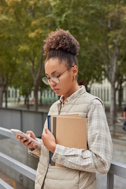 el estudiante revisa la notificación en un teléfono celular moderno lleva blocs de notas lee un mensaje de texto conectado a Internet público al aire libre usa anteojos redondos y ropa informal