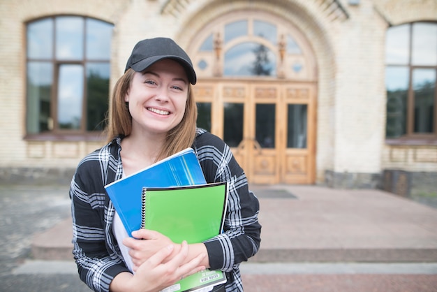 Estudiante en ropa casual con libros y cuadernos en sus manos contra el fondo de un edificio universitario.