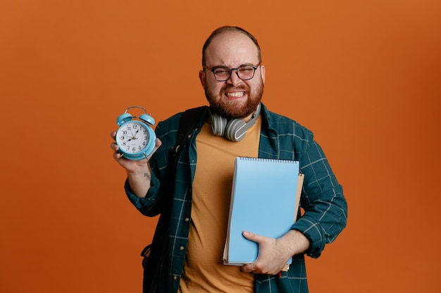 Estudiante con ropa informal y gafas con auriculares sosteniendo cuadernos y reloj despertador mirando a la cámara emocionado sonriendo con expresión molesta de pie sobre fondo naranja