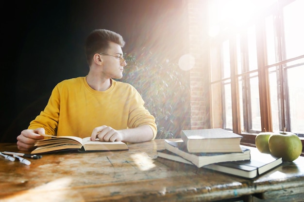 Estudiante rubio con anteojos usando un suéter amarillo mirando a la ventana mientras hace su tarea en la clase