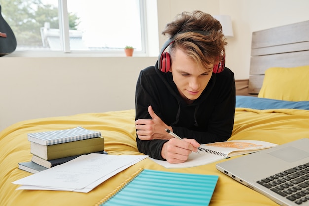 Estudiante de secundaria acostado en la cama en casa y escribiendo en un cuaderno al hacer los deberes para la clase de matemáticas