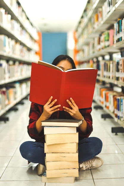 estudiante sentada en el suelo con un montón de libros de educación en la biblioteca cubriendo cara por libro