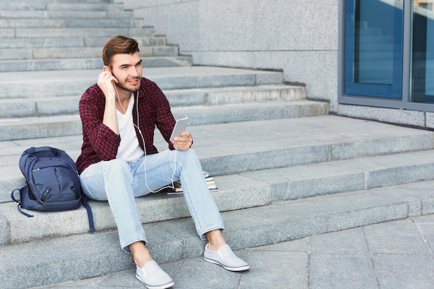 Estudiante sentado en escaleras grises y escuchando música en su smartphone al aire libre, descansando en el campus universitario con espacio para copiar. Concepto de educación, descanso y relax.