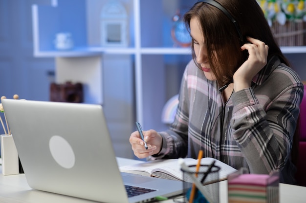 La estudiante seria usa auriculares en línea, estudia en línea con un maestro de internet, aprende un idioma hablando mirando la computadora portátil, una mujer joven enfocada hace una videollamada tutoría escribe notas