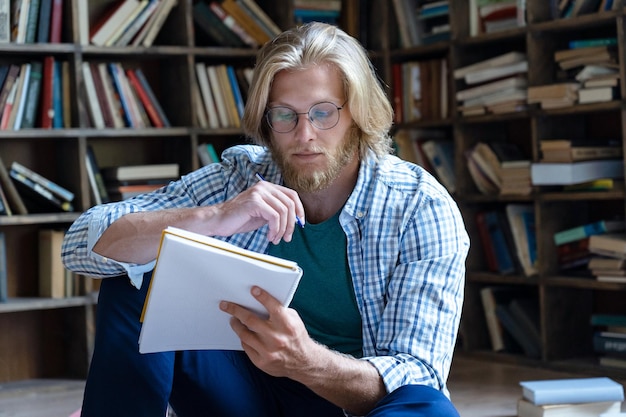 Estudiante serio sosteniendo un libro de trabajo escribiendo un ensayo estudiando sentado en la biblioteca