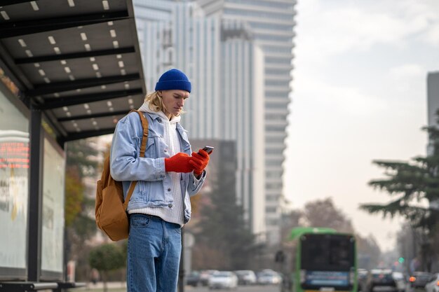 Estudiante de sexo masculino de pie en la parada de autobús mirando la ruta en el mapa de la aplicación de navegador GPS en su teléfono inteligente