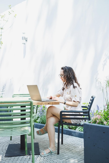 Una estudiante se sienta en la terraza de verano de un café y trabaja en una computadora portátil Una mujer independiente trabaja de forma remota en línea mientras está sentada en un café de verano Trabajo remoto