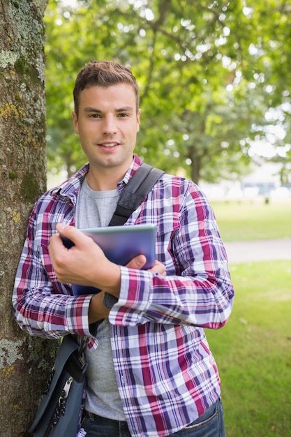 Estudiante sonriente apoyado en el árbol con su tablet pc