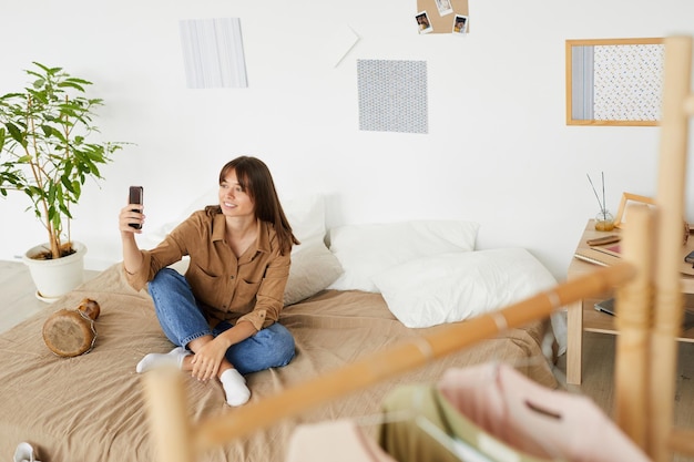 Foto estudiante sonriente con cabello castaño sentada en la cama y con comunicación en línea a través de la aplicación móvil