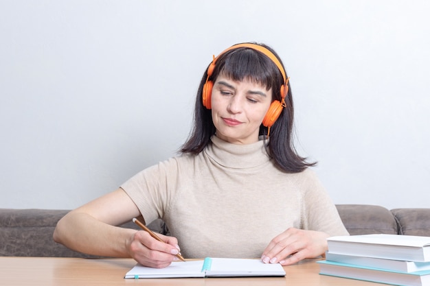 Estudiante sonriente escuchando un curso de audio remoto en auriculares y escribiendo algo en un cuaderno mientras está sentado en la mesa