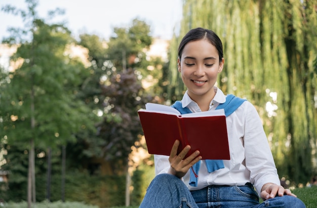 Estudiante sonriente estudiando, aprendiendo el idioma en el parque, leyendo un libro, sentado en el césped. Concepto de educación