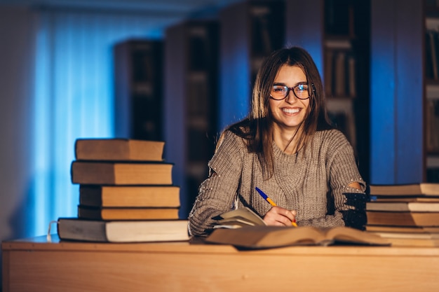 Estudiante sonriente feliz joven en los vidrios que se preparan para el examen. Niña en la noche se sienta a una mesa en la biblioteca con una pila de libros, sonriendo y mirando