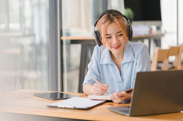 Una estudiante sonriente lleva auriculares inalámbricos estudiando en línea en un café Aprendiendo un idioma escuchando conferencias viendo seminarios web y tomando notas Representa la educación a distancia