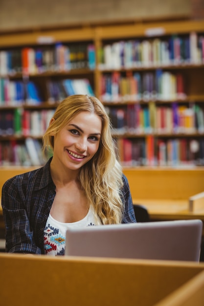 Estudiante sonriente que trabaja mientras que usa su computadora portátil en biblioteca