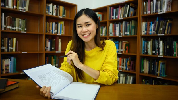 Estudiante sonriente sentada entre estanterías en la biblioteca leyendo libros para estudiar e investigar el concepto de educación