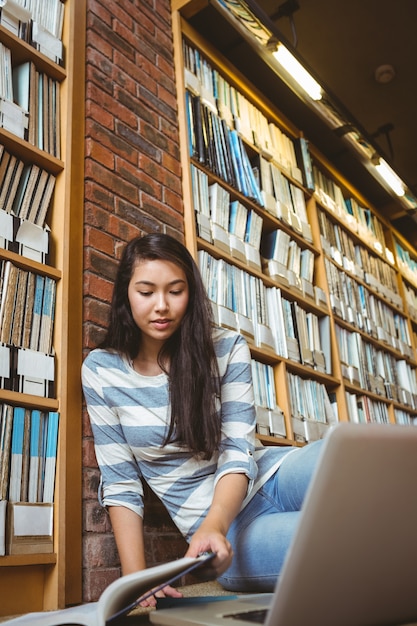 Estudiante sonriente sentada en el piso contra la pared en la biblioteca estudiando con computadora portátil y libros