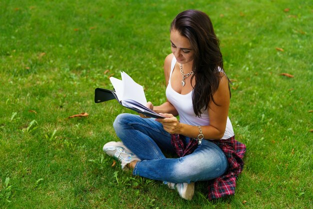 Estudiante sonriente sentado en la hierba con el bloc de notas en la mano, preparándose para los exámenes. Concepto de educación y trabajo remoto. Enfoque selectivo suave. Chica caucásica con una mirada pensativa