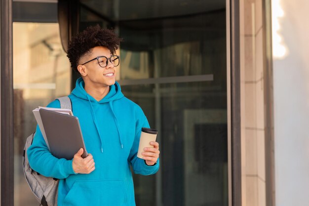 Un estudiante sonriente sosteniendo un café para llevar, una computadora portátil y cuadernos de trabajo afuera.