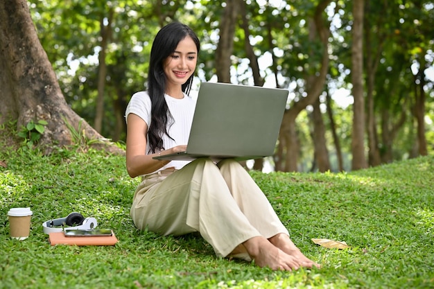 Estudiante universitaria bastante asiática haciendo una tarea en línea en su computadora portátil en el parque de la ciudad