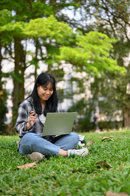 Una estudiante universitaria bastante asiática usando su laptop y sentada en el césped en el parque del campus