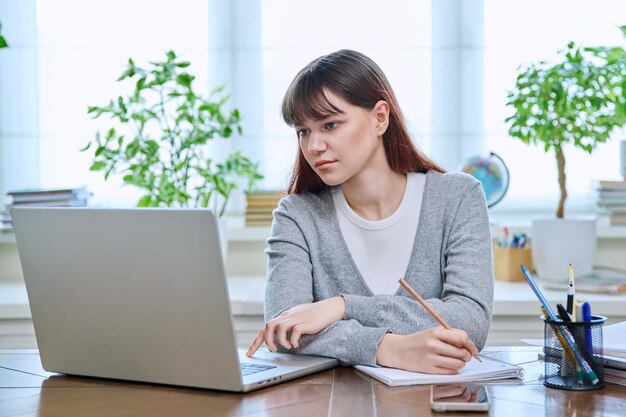 Una estudiante universitaria sentada en un escritorio usando una computadora tomando notas en un cuaderno