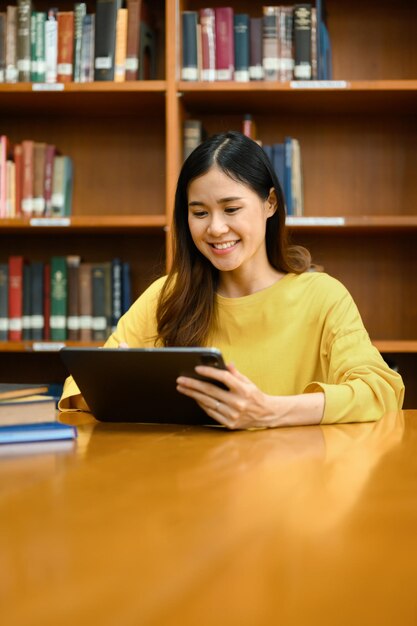 Estudiante universitaria sonriente usando tableta digital investigando lecciones de aprendizaje preparándose para los exámenes en la biblioteca