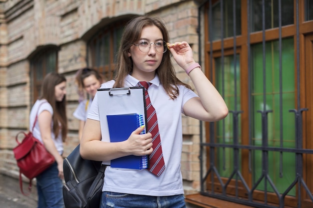 Estudiante universitario de adolescente chica posando al aire libre en camiseta blanca con corbata en gafas. Edificio de ladrillo de fondo, grupo de estudiantes de niñas. Inicio de clases, regreso a la universidad, espacio de copia