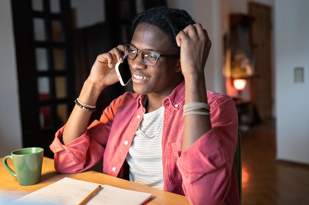 Un estudiante universitario africano sonriente hablando por teléfono mientras estudiaba en casa tomando un descanso del estudio