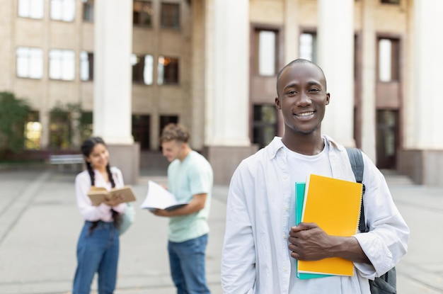 Foto estudiante universitario afroamericano masculino con mochila y libros de trabajo al aire libre posando cerca de la universidad