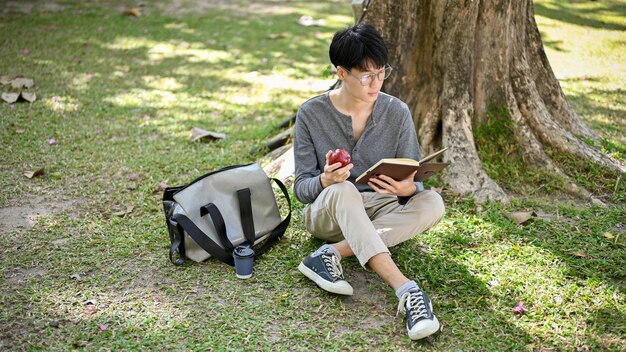 Un estudiante universitario asiático relajado comiendo manzana mientras lee un libro debajo del árbol