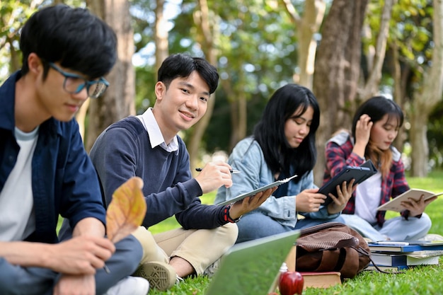 Estudiante universitario asiático usando su tableta mientras está sentado en el parque de la universidad con sus amigos