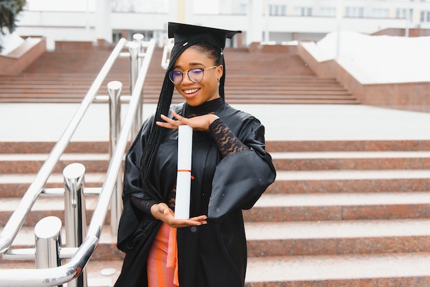 Estudiante universitario bastante africano en toga y birrete de graduación en frente del edificio de la escuela