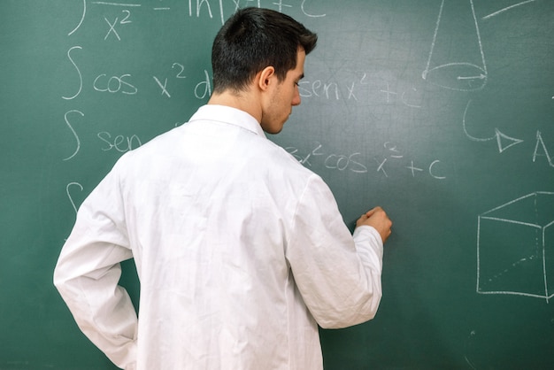 Estudiante universitario en clase de laboratorio, con bata blanca, escribiendo en la pizarra.