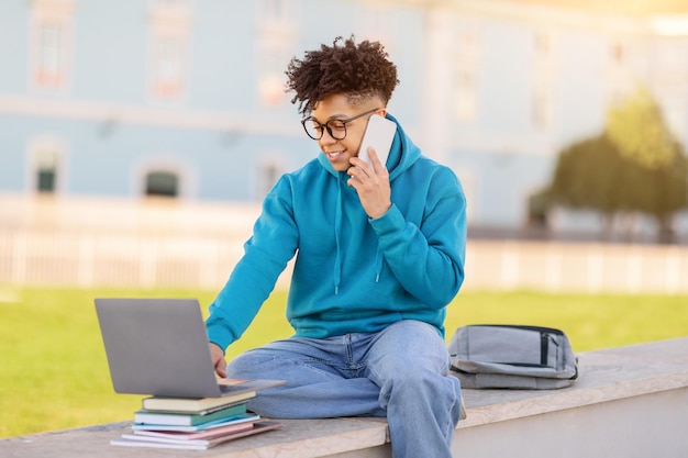 Un estudiante universitario habla por teléfono, navega por la computadora portátil al aire libre.