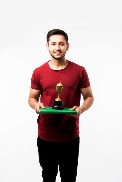 Estudiante universitario indio con libro, leyendo o mostrando la copa de la victoria, premio mientras está de pie aislado sobre fondo blanco.