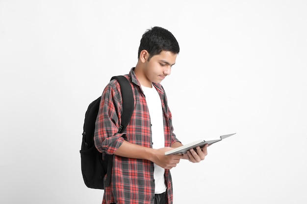 Estudiante universitario indio sosteniendo la mochila y el libro de lectura en la pared blanca