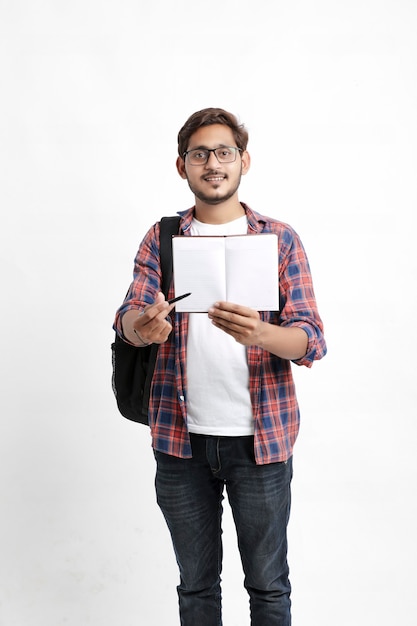 Foto estudiante universitario indio sosteniendo la mochila y mostrando el cuaderno en la pared blanca