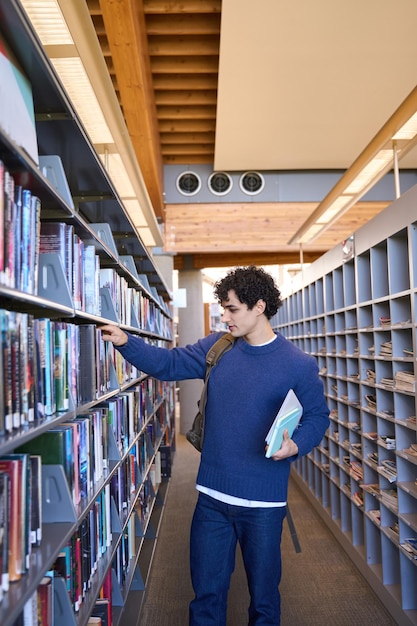 Estudiante universitario inteligente latinoamericano elige un libro junto a estanterías en el campus de la biblioteca