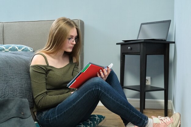 Estudiante universitario joven con libros de texto leyendo y estudiando en casa, educación a distancia. Chica inteligente con gafas sentado en el piso