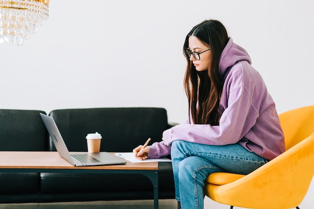 Estudiante universitario de mujer joven caucásica enfocada en anteojos estudiando con computadora portátil, preparándose a distancia para el examen de prueba, escribiendo un ensayo haciendo la tarea en casa, concepto de educación a distancia