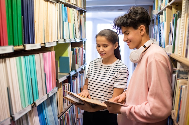 Estudiante universitario con tableta y su compañero de clase mirando a través del libro en la biblioteca mientras está de pie entre estanterías