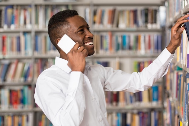 Estudiante varón africano hablando por teléfono en la biblioteca con poca profundidad de campo.