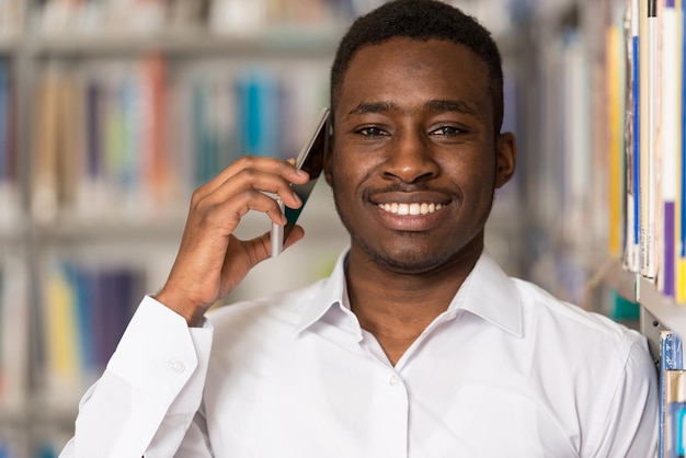 Estudiante varón africano hablando por teléfono en la biblioteca con poca profundidad de campo.