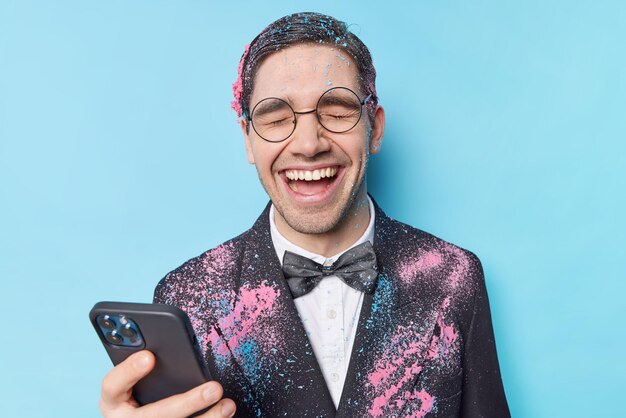 Estudiante varón muy contento disfruta de la fiesta de despedida de la universidad usa espectáculos redondos y traje formal usa el teléfono celular para charlar risas alegremente posa contra el fondo azul Concepto de celebración de la gente