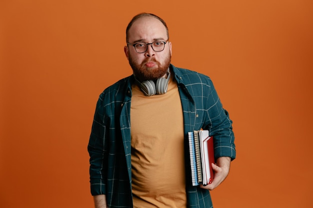 Estudiante vestido con ropa informal con gafas y auriculares sosteniendo cuadernos mirando a la cámara ofendido con una expresión triste de pie sobre un fondo naranja