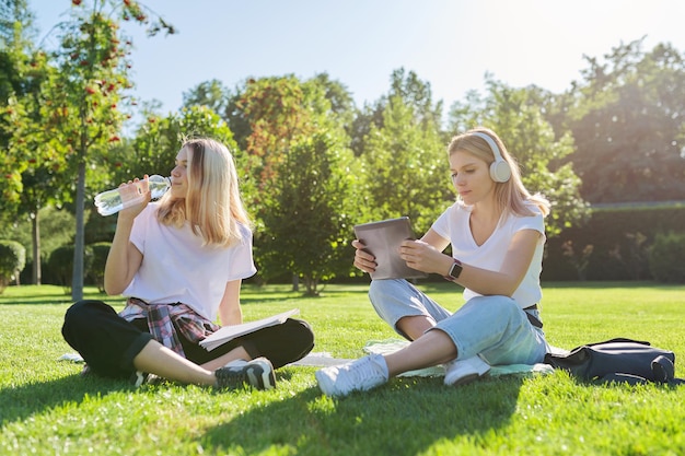 Estudiantes adolescentes niñas sentados en el césped verde en el parque con mochila, libros, tableta digital, botella de agua potable. Universidad, colegio, escuela, educación y conocimiento, estilo de vida adolescente