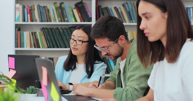 Los estudiantes amistosos se sientan en el escritorio en la escritura de la biblioteca discuten el proyecto colaborativo que se prepara para los exámenes de la escuela secundaria Concepto de estudio de educación universitaria